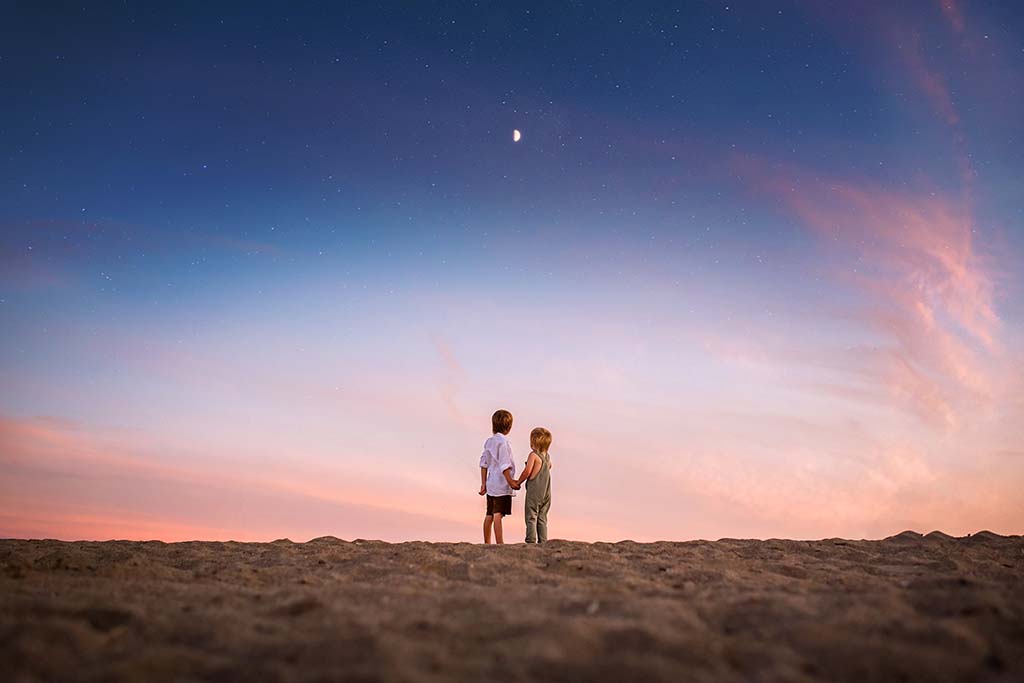 2 children holding hands and staring at the night sky with the moon and stars in the backround